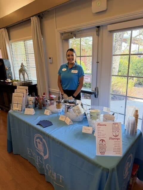 Smiling staff member in a blue polo stands behind a promotional table covered with a light‑blue cloth, displaying samples and handouts indoors.